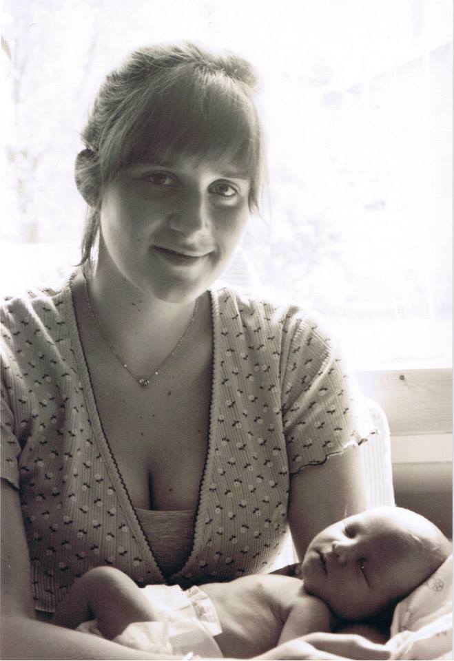 A young mother smiles at the camera while holding her sleeping newborn on her lap.