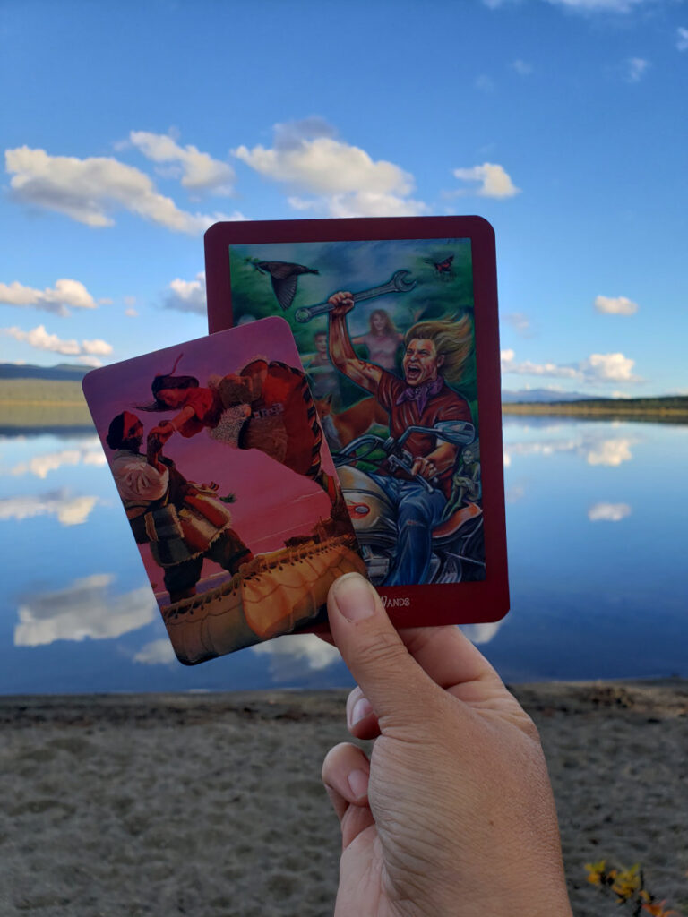 Tarot and oracle cards are held up in front of a glassy northern lake reflecting clouds.