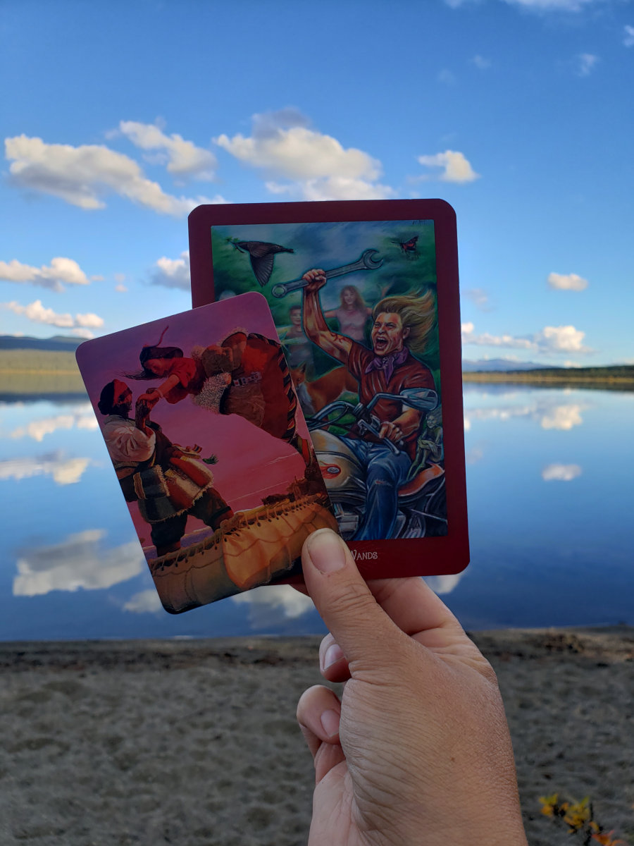 Tarot and oracle cards are held up in front of a glassy northern lake reflecting clouds.